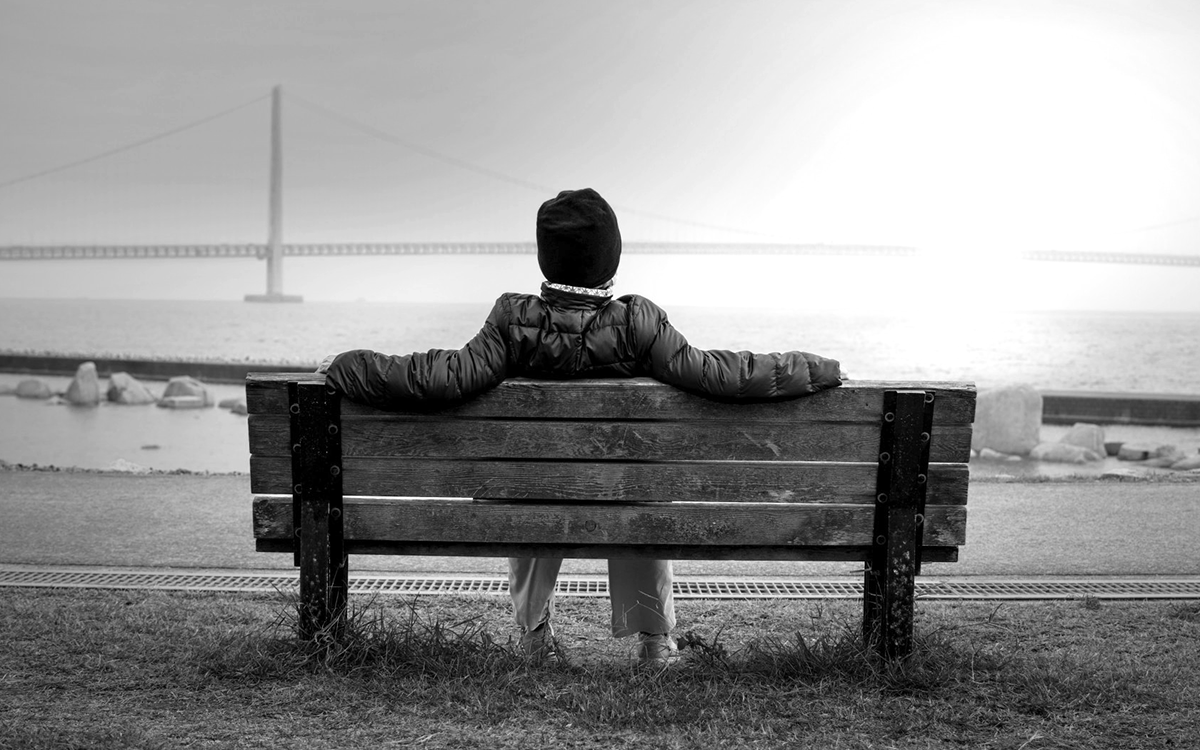 A person sitting on a weathered wooden bench beside a calm waterfront.