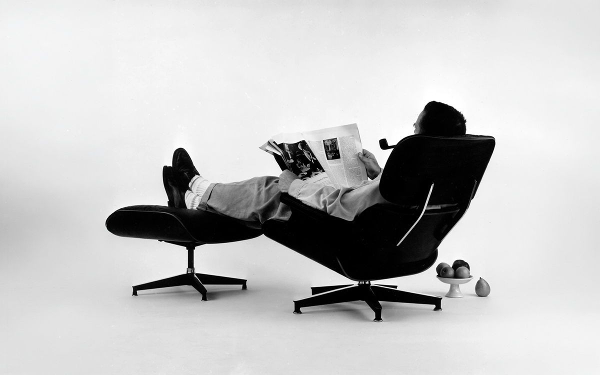 A man relaxing in an Eames lounge chair with reading a newspaper and smoking a pipe.